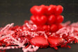 A red heart-shaped candle sits in the background against a dark surface, slightly out of focus. In the foreground, two heart-shaped chocolates wrapped in shiny red foil rest among red and pink crinkled paper shreds. The glossy textures of the candle and chocolates reflect soft light, contrasting with the matte paper decorations. The composition highlights the rich red tones and smooth surfaces. Photography.