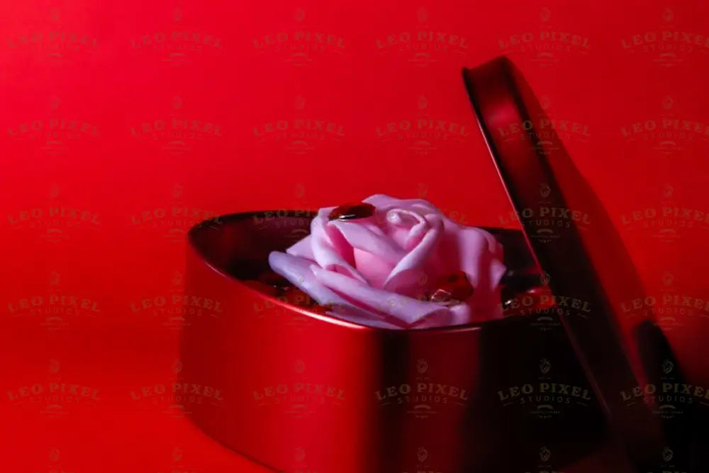 A red heart-shaped box sits slightly open, revealing a delicate pink artificial rose placed inside. The rose is surrounded by small, shiny red gems. The vivid red background complements the box and enhances the soft tones of the rose. The glossy texture of the box and the smooth petals create a visually striking composition. Photography.