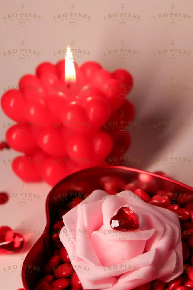 A red heart-shaped metal box is filled with glossy red hearts, topped with a soft pink rose with a faceted red heart gem on its petals. In the softly blurred background, a red bubble candle with heart details glows warmly, casting soft shadows. Scattered red crystal hearts add texture, while the warm lighting highlights the rich colors and smooth surfaces. Photography.