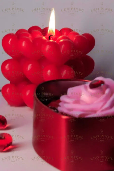 A red heart-shaped metal box is filled with glossy red hearts, topped with a soft pink rose with a faceted red heart gem on its petals. In the background, a red bubble candle with heart details glows warmly, casting soft shadows. Scattered red crystal hearts add texture, while the warm lighting highlights the rich colors and smooth surfaces. Photography.