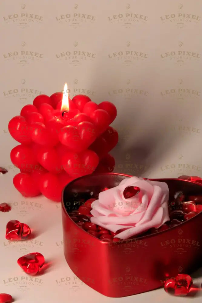 A red heart-shaped metal box is filled with glossy red hearts, topped with a soft pink rose with a faceted red heart gem on its petals. In the background, a red bubble candle with heart details glows warmly, casting soft shadows. Scattered red crystal hearts add texture, while the warm lighting highlights the rich colors and smooth surfaces.