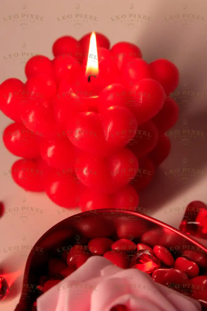 A red heart-shaped metal box is filled with glossy red hearts, topped with a soft pink rose with a faceted red heart gem on its petals. In the background, a red bubble candle with heart details glows warmly, casting soft shadows. Scattered red crystal hearts add texture, while the warm lighting highlights the rich colors and smooth surfaces. Photography.