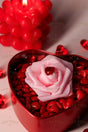 A red heart-shaped metal box is filled with glossy red hearts, topped with a soft pink rose with a faceted red heart gem on its petals. In the softly blurred background, a red bubble candle with heart details glows warmly, casting soft shadows. Scattered red crystal hearts add texture, while the warm lighting highlights the rich colors and smooth surfaces. Photography.