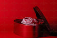 A red heart-shaped box sits slightly open, revealing a delicate pink artificial rose placed inside. The rose is surrounded by small, shiny red gems. The vivid red background complements the box and enhances the soft tones of the rose. The glossy texture of the box and the smooth petals create a visually striking composition. Photography.