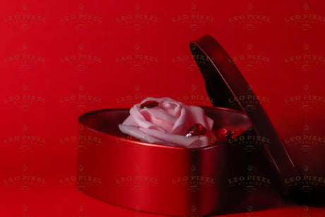 A red heart-shaped box sits slightly open, revealing a delicate pink artificial rose placed inside. The rose is surrounded by small, shiny red gems. The vivid red background complements the box and enhances the soft tones of the rose. The glossy texture of the box and the smooth petals create a visually striking composition. Photography.