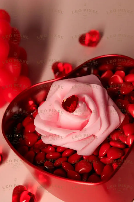 A red heart-shaped metal box is filled with glossy red candy-coated chocolates and a delicate pink rose. A small, faceted red heart-shaped gem rests on the petals. In the softly blurred background, a pink bubble-shaped candle with heart details and scattered red heart decorations add depth. The warm, diffused lighting enhances the soft textures and rich red hues. Photography.