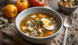 A bowl of hearty soup with a clear, golden broth, containing chunks of tender vegetables like potatoes, carrots, and celery. A halved boiled egg rests on top, garnished with fresh basil leaves. The soup is accompanied by soft bread-like pieces. Vibrant yellow bell peppers and tomatoes are in the background, with a spoon placed nearby on a fringed napkin on a wooden surface. Ai generated. Photography style.