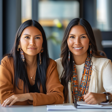 Image of two Indigenous women seated side by side at a desk in a modern office setting. They wear elegant attire complemented by traditional beaded earrings and layered necklaces. Both women have long, dark hair and are smiling confidently, with notebooks and pens placed in front of them on the table.