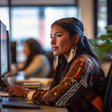 An image of a Native American woman working at a computer in a modern office. She wears a patterned brown top and large feather earrings, with her dark hair styled back. She looks attentively at the monitor, one hand on the mouse. The blurred background shows another woman at a computer and a softly lit workspace.