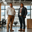 Two Indigenous men stand confidently in a modern office space with floor-to-ceiling windows. One wears a white shirt and tan pants, while the other pairs a dark blazer with a beaded medallion necklace, white shirt, and dark trousers. Both men have their hands in their pockets and maintain a composed, serious expression.