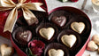 A beautifully arranged heart-shaped gift box holds exquisite gold and dark chocolate hearts, each delicately inscribed with romantic cursive details. Nestled in dark paper wrappers, the chocolates gleam under soft light, exuding elegance. A rich golden ribbon adorns the box, accompanied by a single red rose, creating a perfect, indulgent symbol of love and affection. Ai generated image.