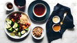 A well-balanced meal arrangement on a clean, white surface features a large plate of steamed broccoli florets, colorful carrots, grapes, and shredded vegetables, accompanied by a small dish of granola. Two deep bowls hold rich, amber-colored soup or tea, while a cup of dark liquid adds contrast. A dark cloth napkin with a golden fork completes the refined presentation. Ai generated. Photography style.