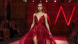 A model walks confidently down the runway in a flowing, vibrant red gown with a deep V neckline and spaghetti straps. The gown's fabric shimmers under the warm lights, with a high slit revealing her stride. Her long, wavy hair complements the elegance of her look. The blurred audience in the background claps, while red neon accents enhance the stage atmosphere. Ai generated. Photography style.