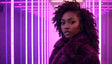 A stunning woman with defined features and natural curly hair stands against a backdrop of vivid pink and purple neon lights. Her confident gaze is framed by the luxurious texture of her deep violet fur coat. The alternating vertical light bars create a sleek, futuristic ambiance that contrasts beautifully with the softness of her attire, enhancing the image's modern elegance. Ai generated. Photography style.