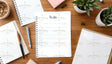 A wooden desk holds multiple spiral-bound notebooks, with one open in the center displaying a "To do" list. A black and brown pen rests on a closed planner, while a blue pen lies nearby. Small potted succulents are placed around the workspace. A bowl with granola sits in the top left, and a pastel pink book is near the top. The layout is neat and well-balanced, with soft natural lighting. Ai generated. Photography style.