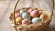 The image shows a wicker basket filled with pastel-colored Easter eggs. Each egg is decorated with delicate patterns, including flowers, polka dots, and zigzag designs. The eggs are in soft shades of pink, yellow, blue, and beige, resting on straw inside the basket. The background is a smooth, light wooden surface, complementing the soft and natural feel of the composition. Ai generated. Photography style.