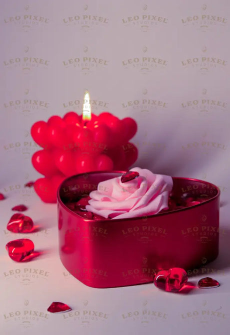 A heart-shaped red metal box filled with glossy red candy-coated hearts holds a soft pink artificial rose with a faceted red heart gem on its petals. Behind it, a glowing red bubble candle with heart-shaped details casts a warm light. Scattered red crystal hearts add texture. The soft lighting highlights the rich red and pink tones against a neutral background. Photography.