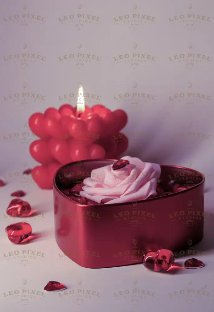 A heart-shaped red metal box filled with glossy red candy-coated hearts holds a soft pink artificial rose with a faceted red heart gem on its petals. Behind it, a glowing red bubble candle with heart-shaped details casts a warm light. Scattered red crystal hearts add texture. The soft lighting highlights the rich red and pink tones against a neutral background. Photography.