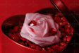 A close-up of a red heart-shaped box holds a soft pink artificial rose at its center, surrounded by glossy red heart-shaped gems and small red pebbles. The smooth texture of the rose contrasts with the shiny gems, creating a striking composition against the vivid red background. Photography style.