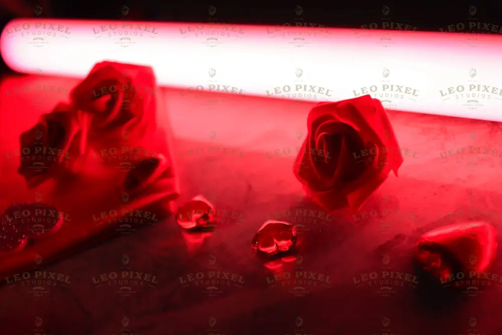 A vivid red light illuminates the scene, highlighting artificial red roses on a flat surface. Two small glass heart-shaped decorations and a folded red napkin add texture. The soft glow contrasts with shadows, enhancing the roses' delicate details. The background blends into the vibrant red ambiance, creating a dramatic and striking visual. Photography.