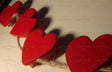 A close-up shot of a string of red wooden hearts attached to a rustic twine rope, casting soft shadows on a white wall. The hearts are arranged in a row, with some appearing slightly out of focus. Warm lighting enhances the deep red tones, creating a cozy and romantic visual effect. The texture of the rope contrasts with the smooth, glossy surface of the hearts. Photography.