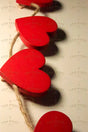 A close-up shot of a string of red wooden hearts attached to a rustic twine rope, casting soft shadows on a white wall. The hearts are arranged in a row, with some appearing slightly out of focus. Warm lighting enhances the deep red tones, creating a cozy and romantic visual effect. The texture of the rope contrasts with the smooth, glossy surface of the hearts. photography.