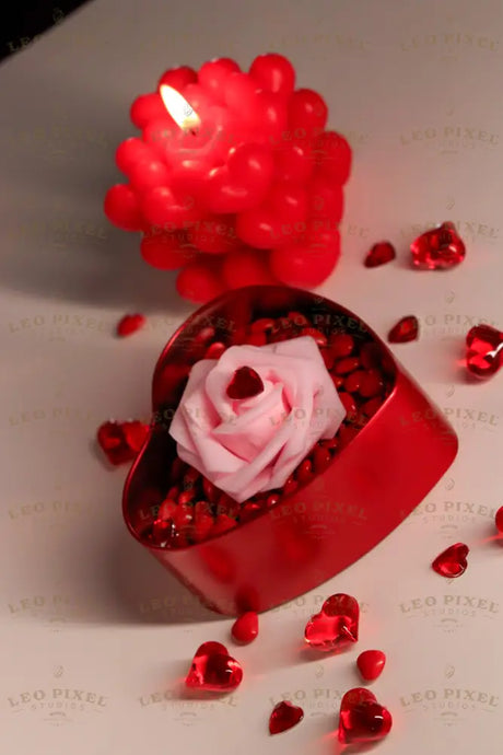 A red heart-shaped tin filled with glossy red candy holds a delicate pink artificial rose with a red gemstone at its center. Scattered around are small red glass hearts and candy pieces. In the background, a red bubble candle with a lit flame glows softly. The scene is set against a neutral background, highlighting the deep red hues and the textures of the objects. The lighting casts soft shadows. Photography.