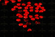 A collection of small, smooth, red heart-shaped beads is scattered across a matte black surface. The glossy beads catch light, creating a subtle shine. Among the beads are a few translucent red gem-like hearts, adding texture and contrast to the arrangement. The dark background enhances the vividness of the red tones, creating a striking visual effect. Photography.