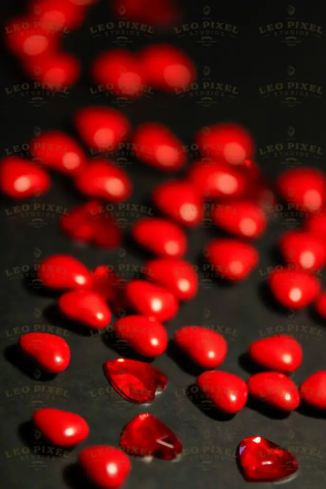 A collection of small, smooth, red heart-shaped beads is scattered across a matte black surface. The glossy beads catch light, creating a subtle shine. Among the beads are a few translucent red gem-like hearts, adding texture and contrast to the arrangement. The dark background enhances the vividness of the red tones, creating a striking visual effect. Photography.