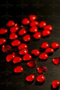 A collection of small, smooth, red heart-shaped beads is scattered across a matte black surface. The glossy beads catch light, creating a subtle shine. Among the beads are a few translucent red gem-like hearts, adding texture and contrast to the arrangement. The dark background enhances the vividness of the red tones, creating a striking visual effect. Photography.