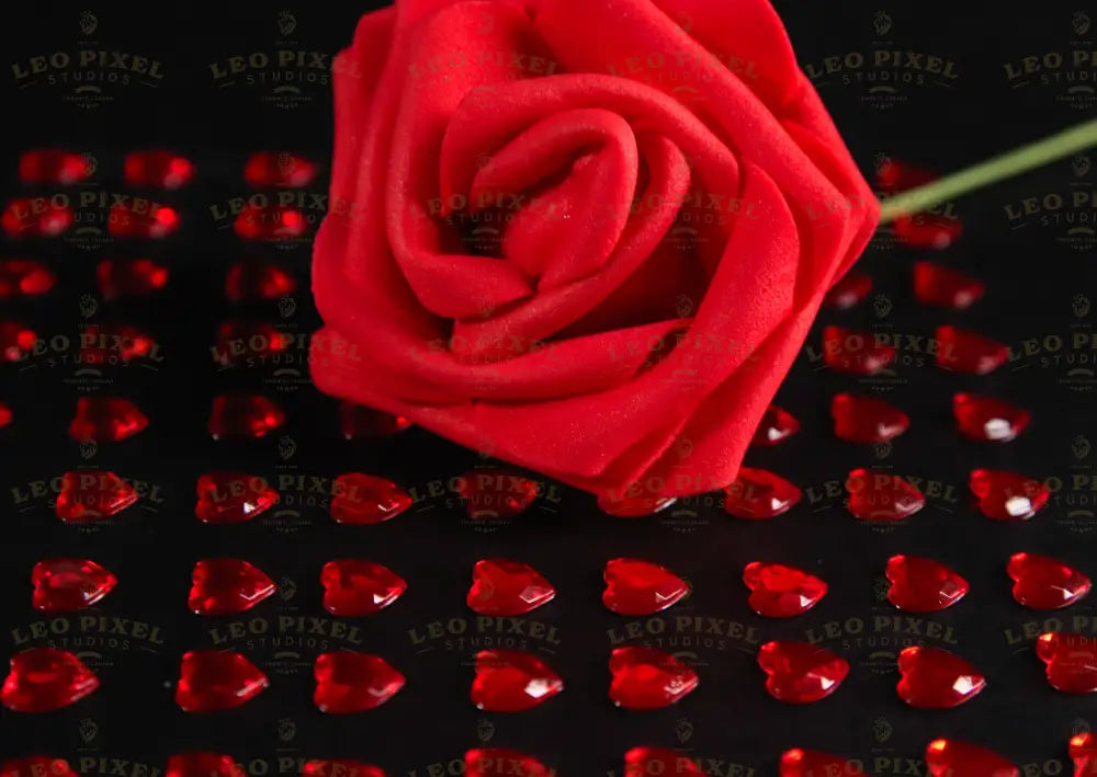 A close-up of a red artificial rose placed on a black surface, surrounded by small, glossy red heart-shaped gems arranged in a grid pattern. The vibrant red contrasts with the dark background, highlighting the details of the fabric petals and the reflective surfaces of the gems. The soft lighting enhances the depth and texture, creating subtle shadows that add dimension to the composition. Photography.