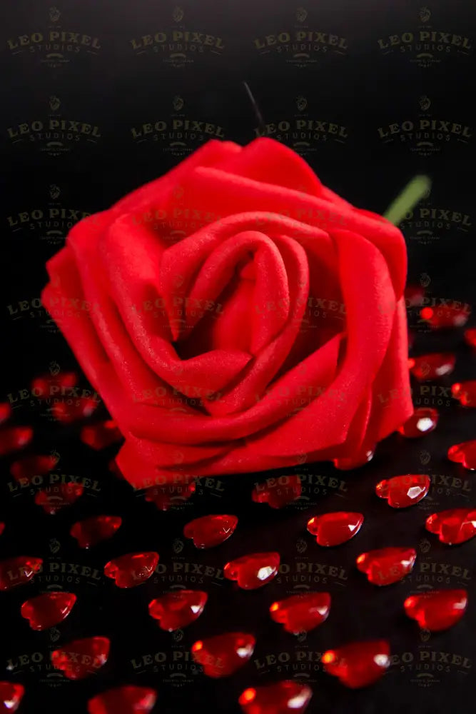 A close-up of a red artificial rose placed on a black surface, surrounded by small, glossy red heart-shaped gems arranged in a grid pattern. The vibrant red contrasts with the dark background, highlighting the details of the fabric petals and the reflective surfaces of the gems. The soft lighting enhances the depth and texture, creating subtle shadows that add dimension to the composition. Photography.