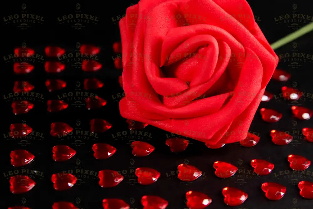 A close-up of a red artificial rose placed on a black surface, surrounded by small, glossy red heart-shaped gems arranged in a grid pattern. The vibrant red contrasts with the dark background, highlighting the details of the fabric petals and the reflective surfaces of the gems. The soft lighting enhances the depth and texture, creating subtle shadows that add dimension to the composition. Photography.