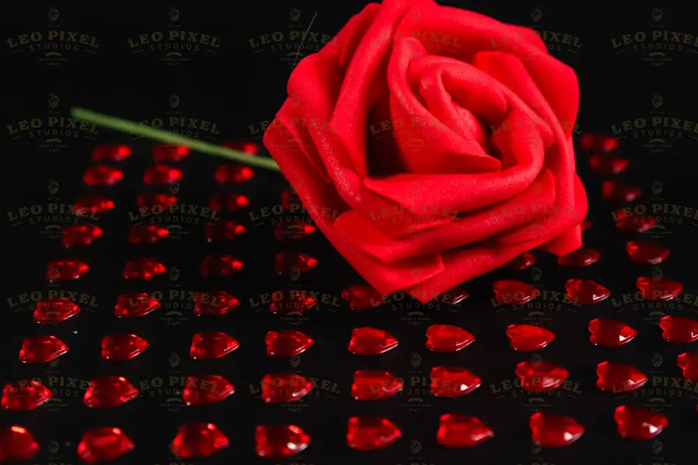 A close-up of a red artificial rose placed on a black surface, surrounded by small, glossy red heart-shaped gems arranged in a grid pattern. The vibrant red contrasts with the dark background, highlighting the details of the fabric petals and the reflective surfaces of the gems. The soft lighting enhances the depth and texture, creating subtle shadows that add dimension to the composition. Photography.