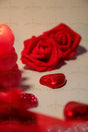 A close-up photograph features two red artificial roses with velvety petals lying on a smooth surface. In the foreground, a shiny red heart-shaped chocolate is wrapped in metallic foil, casting a soft shadow. A blurred heart candle appears on the left, partially visible. The background has a gentle blur, emphasizing the rich red hues and soft lighting, creating a warm and romantic feel. Photography style.