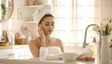 A serene woman with a towel wrapped around her head and shoulders stands in a bright bathroom, eyes closed and hand touching her cheek. The minimalist space features a white countertop, a faucet, fresh towels, and a vase of flowers. The background includes soft lighting, shelves with neatly arranged items, and a round mirror, creating a clean and calming atmosphere. Ai generated. Photography style.