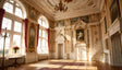 A luminous Rococo gallery showcases grand arched windows framed by red drapes, flooding the room with sunlight. Delicate gold details embellish cream walls adorned with portraits and reliefs. Ornate chandeliers hang from an intricately decorated ceiling. A curved staircase complements the refined interior, while a vase of vibrant flowers atop a classic table adds a graceful touch. Ai generated. Photography style.