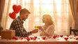 A couple shares a tender moment, clinking glasses in soft golden sunlight. Red heart-shaped balloons and scattered rose petals enhance the romantic atmosphere. A gift box rests nearby, symbolizing affection and surprise. The warm glow filtering through the curtains highlights their connection, creating a scene that embodies love, joy, and the beauty of shared moments. Ai generated. Photography style.