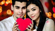 A vibrant, close-up cartoon-style image of a loving couple holding a bright red heart together. The man, with short dark hair and a white shirt, smiles warmly while the woman, with long black hair and soft features, gazes tenderly. The background glows with soft, blurred lights and warm red tones, creating a cozy, intimate atmosphere that radiates love and joy. Ai generated image. Photography style.