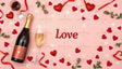 A flat lay image on a pink background showcases a champagne bottle, wine glasses, red hearts, ribbons, small white flowers, and fresh green sprigs. The word "Love" appears in bold red letters, surrounded by decorative elements that create a romantic and celebratory mood. Ai generated. Flat lay photography style