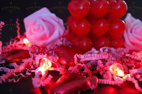 A red heart-shaped cube candle sits among soft pink roses on a dark surface. The scene is decorated with heart-shaped foil-wrapped chocolates, glowing fairy lights, and shredded paper. The blurred background enhances the glow of the lights, creating a cozy and romantic feel. The deep reds and soft pinks stand out against the dark setting, making it perfect for Valentine's Day themes. Photography.