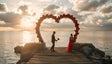 A couple stands on a serene wooden pier at sunset, framed by a large heart-shaped arch adorned with vibrant red roses. The golden sun sets over the calm ocean, casting a warm glow and reflecting on the water’s surface. The man, dressed in a suit, holds a rose bouquet as he faces a woman in an elegant red gown. Gentle clouds float overhead, enhancing the romantic and timeless atmosphere. Ai generated image. Photography style.