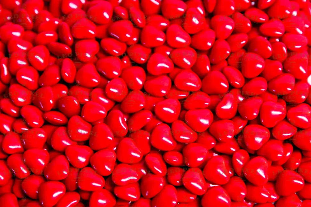 A close-up shot of glossy red heart-shaped candies fills the entire frame. The candies are packed tightly together, creating a textured surface with soft highlights reflecting off their smooth coating. The vibrant red color stands out, making the image bright and eye-catching. The focus is sharp, capturing each detail, while the lighting enhances the glossy look, giving a warm and inviting feel. Photography.