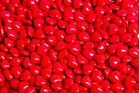 A close-up shot of glossy red heart-shaped candies fills the entire frame. The candies are packed tightly together, creating a textured surface with soft highlights reflecting off their smooth coating. The vibrant red color stands out, making the image bright and eye-catching. The focus is sharp, capturing each detail, while the lighting enhances the glossy look, giving a warm and inviting feel. Photography.