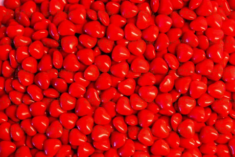 A close-up shot of glossy red heart-shaped candies fills the entire frame. The candies are packed tightly together, creating a textured surface with soft highlights reflecting off their smooth coating. The vibrant red color stands out, making the image bright and eye-catching. The focus is sharp, capturing each detail, while the lighting enhances the glossy look, giving a warm and inviting feel. Photography.