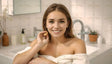 A young woman with long, light brown hair smiles warmly, holding a skincare product near her face. She is wrapped in a white towel, standing in a bright, modern bathroom. The background features a white sink, potted plant, soap dispenser, and subtle decor. The setting is clean and inviting, with soft natural lighting enhancing the serene and fresh atmosphere. Ai generated. Photography style.