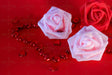 A red background holds two soft pink roses and one deep red rose, each with delicate petal folds. The roses are surrounded by glossy red heart-shaped beads forming a chain, reflecting light. Scattered across the surface, smaller heart gems add texture and shine. The rich red tones contrast with the soft pastel hues, creating a balanced composition with a focus on romance and elegance. Photography. 