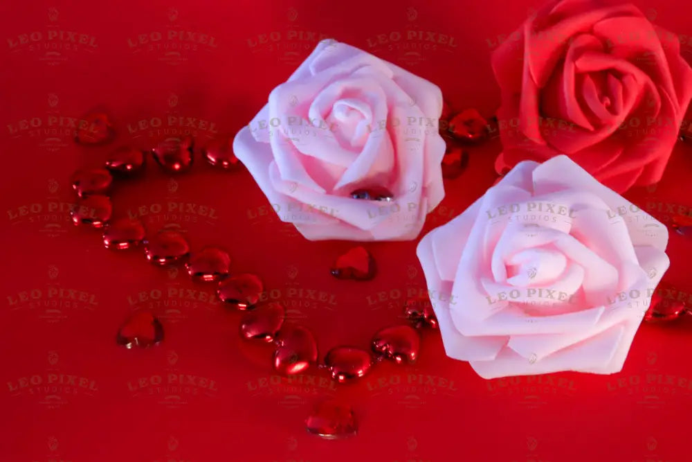 A red background holds two soft pink roses and one deep red rose, each with delicate petal folds. The roses are surrounded by glossy red heart-shaped beads forming a chain, reflecting light. Scattered across the surface, smaller heart gems add texture and shine. The rich red tones contrast with the soft pastel hues, creating a balanced composition with a focus on romance and elegance. Photography. 