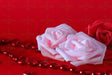 Three roses, two in soft pink and one in deep red, rest against a red background. Small red heart-shaped gems are scattered around, reflecting light. The petals have a velvety texture, and the green stem of the red rose adds contrast. The smooth backdrop enhances the warm tones, while the gems create a delicate sparkle in the composition. Photography.