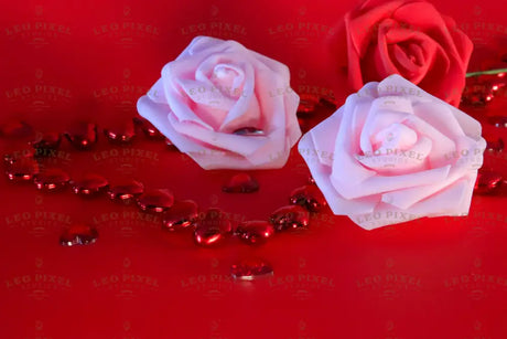 Three roses, two in soft pink and one in deep red, rest against a red background. Small red heart-shaped gems are scattered around, reflecting light. The petals have a velvety texture, and the green stem of the red rose adds contrast. The smooth backdrop enhances the warm tones, while the gems create a delicate sparkle in the composition.